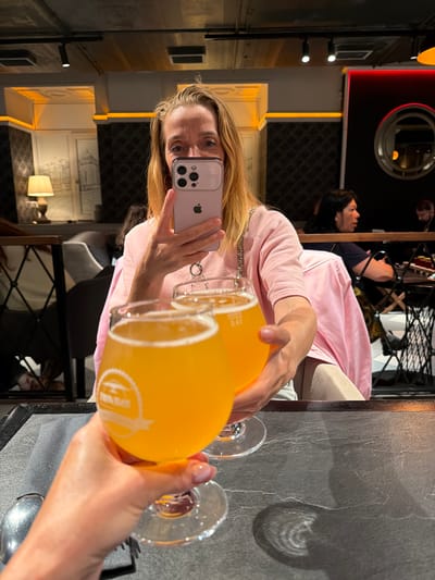 The image shows a woman holding a phone and two glasses of yellow liquid, likely beer or juice, in a dimly lit bar or restaurant. The woman, with blonde hair, is taking a selfie with her phone, which is pink and appears to be an iPhone. She is wearing a light pink t-shirt and a silver chain necklace. In the background, two other people are visible, one wearing a dark shirt and the other a pink top. The setting appears to be a modern establishment with dark grey textured walls and decorative lighting. Reflections in the background suggest mirrors and intricate architectural details, possibly indicating a historical building. One of the glasses has a white logo with text. The overall mood is casual and relaxed, suggesting a social gathering or a meal out. The time of day is likely evening or night due to the interior lighting. The location is specified as Saint Petersburg, Russia.