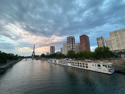 A wide-angle, eye-level shot captures the Seine River in Paris, France, under a dramatic, cloudy sky at dusk. The Eiffel Tower is visible in the distance on the left, with a bridge spanning the river in front of it. A large, white river cruise ship is docked along the right bank, its numerous windows reflecting the dim light. Behind the ship, a line of modern high-rise buildings forms the city's skyline. Trees line the riverbanks, and a few individuals can be seen walking along the promenade next to the ship. The water of the Seine is dark and slightly rippled, reflecting the muted colors of the sky and city lights.