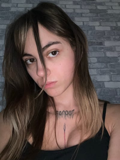 A woman with dark brown hair and bangs is captured in a close-up portrait. She is wearing a black tank top and has a tattoo on her chest and neck. The background consists of a gray brick wall. The image does not provide any information about the location in Moscow, Russia.