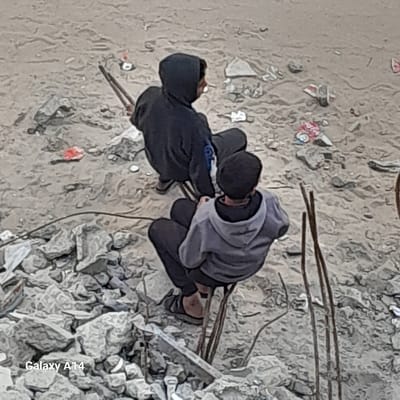An overhead view captures two individuals seated amidst a debris-strewn landscape in Khan Yunis, Palestinian Territory. The ground is a mix of light-colored soil, sand, and abundant rubble, primarily fragmented concrete and rocks. The individual in the foreground, facing away from the camera, wears a light gray hooded sweatshirt, dark pants, and dark flip-flops, crouching on exposed, bent metal rods. Behind and to their left, another individual, wearing a dark hooded sweatshirt and dark pants, holds a long, dark, two-pronged gripping tool. This individual is also seated or crouched on similar metal rods. Both individuals are oriented towards the upper right portion of the frame. The immediate environment includes scattered refuse, such as fragments of red and white plastic wrappers, alongside numerous pieces of construction debris and protruding metal rebar. The background maintains a consistent appearance of disturbed earth and structural wreckage.
