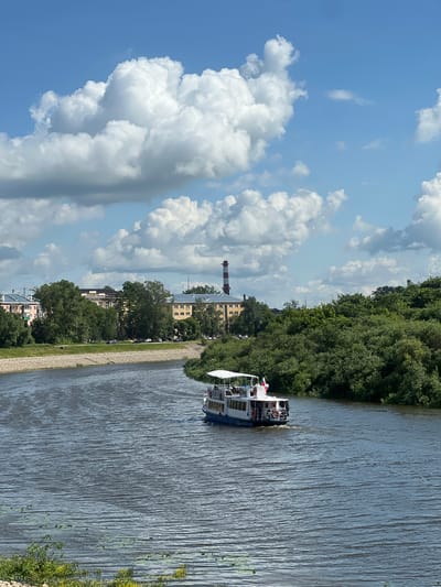 The image shows a wide, dark blue river flowing through a city under a bright blue sky dotted with fluffy white clouds. A white tour boat with a red and white striped flag is cruising on the river. Along the left bank of the river, there is a stone embankment, a grassy area, and several multi-story buildings. On the right bank, there is a dense green forest. In the background, a tall red and white striped smokestack is visible. The scene depicts a calm day in Vologda, Russia, likely during the summer.