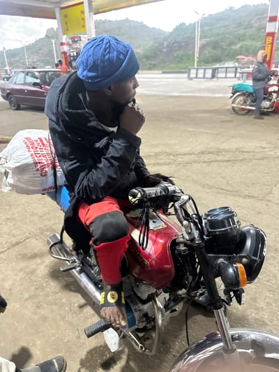 A person wearing a blue knitted beanie and a black jacket over a white shirt sits on a red motorcycle at a gas station. The person is also wearing red pants with black trim and a black wristband with yellow lettering. A large white bag is strapped to the back of the motorcycle. In the background, a red car is parked, and other motorcycles are visible. The scene appears to be an outdoor setting, possibly in Gura Top, Nigeria, given the presence of the gas station and motorcycles. The lighting suggests it may be overcast or dusk.