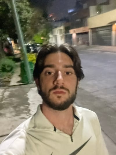 A selfie taken at night on a street in Huixquilucan de Degollado, Mexico shows a young man with dark hair and a beard looking directly at the camera. He is wearing a light-colored Nike jacket with a black collar and a large black Nike logo on the right side of his chest. The street appears to be paved and wet, reflecting the lights of the surrounding buildings and trees. The background shows houses and streetlights, indicating an urban or suburban environment. The overall lighting suggests it is nighttime, and the focus is on the man's face and upper body.