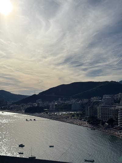 The image shows a wide, aerial view of the coastline in Rafailovići, Montenegro. The sun is visible in the upper left corner, casting a bright glare on the water. The sea is calm, with several boats and yachts anchored near the shore. The beach is lined with many people, and there are numerous buildings and hotels situated along the coastline, backing onto lush green hills. The sky is partly cloudy, with wispy cirrus clouds and a faint contrail from an aircraft. The scene depicts a bustling coastal resort town.