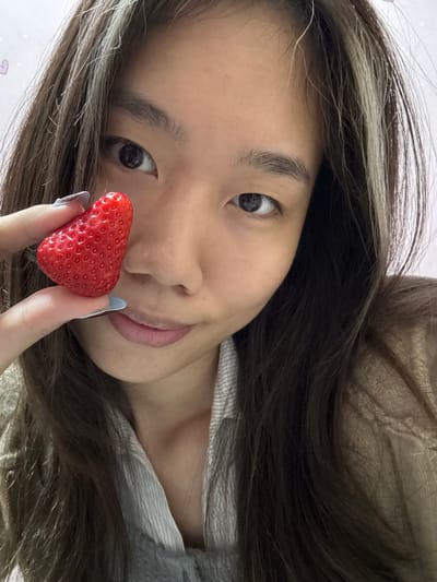 A young woman with long, dark hair and light brown highlights holds a strawberry in front of her face. Her fingernails are painted a pale blue. The background is out of focus but appears to be a light pink surface. The scene is angled as if taken by a selfie. There is no specific evidence indicating the image was taken in Shapingba District, China.