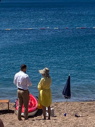 The image shows a man and a woman standing on a sandy beach facing the turquoise water in Petrovac Centar, Montenegro. The man, wearing a white long-sleeved shirt and khaki pants, stands with his back to the camera. The woman, wearing a wide-brimmed straw hat and a yellow dress, stands facing the man. Between them is a bright red kayak with white trim and a black paddle. To the right of the woman, a closed dark blue umbrella is planted in the sand. In the background, buoys mark a section of the sea. A small wooden table is visible on the left side of the frame. The scene suggests a relaxed beach setting.