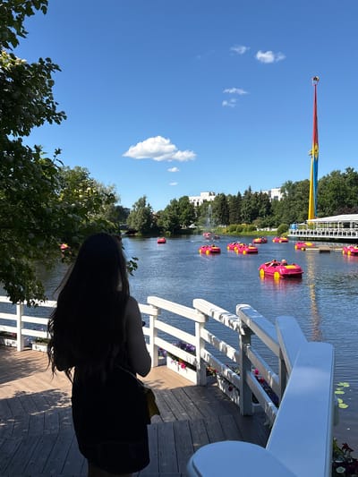 A woman stands on a wooden deck overlooking a lake filled with pink paddle boats shaped like cars. A tall, colorful amusement ride with a red, yellow, and blue tower dominates the skyline. Trees line the shore, with buildings and a fountain visible in the distance. The sky is bright blue with a few white clouds. The scene appears to be located in a park or amusement area in Saint Petersburg, Russia.