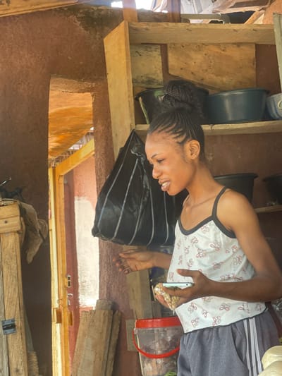 A young woman with braided hair and a white patterned top is smiling and looking down at a phone in her hand. She is holding a small bag of what appears to be peanuts. She is in an indoor setting with wooden shelves and structures visible. The overall scene suggests a casual, possibly domestic environment. The lighting appears to be natural, coming from an unseen source. The woman's expression is happy and engaged. The background includes various containers, possibly buckets or tubs, and some discarded materials, suggesting a functional or storage area. The presence of wooden planks and rough surfaces indicates a potentially rustic or unfinished building. There is no visible text in the image. The time of day and weather are not discernible.