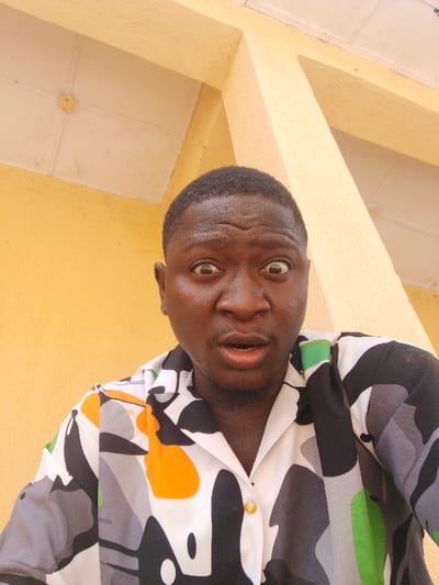 The image displays an adult male, facing the camera directly in a close-up, chest-up shot. He has short, dark hair and exhibits an exaggerated expression characterized by wide eyes and an open mouth. He wears a short-sleeved, collared button-down shirt featuring an abstract pattern in white, black, various shades of grey, bright orange, and green; the top button remains unfastened. The background consists of a solid yellow wall. Above the subject, a white ceiling or soffit is visible, containing a circular fixture in the upper left. A light yellow structural column or beam is present in the upper right background. The specific location within Akwanga, Nigeria, cannot be determined from the visual information provided.