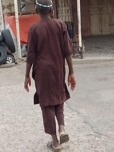The image shows a person from behind, walking away from the camera on a dirt road in Arawa, Nigeria. The person is wearing a long, maroon tunic and matching pants, common attire in the region. They also wear a beaded headband, the beads appearing to be blue and white. On their feet, they have simple, dark-colored flip-flops.
The setting appears to be an outdoor area with some structures in the background. To the left, there are stacks of tires and a reddish-orange tarp, possibly indicating a storage or work area. A white vertical pole, likely part of a makeshift structure, is also visible. Further back, there are buildings with doorways and what might be shelves or stacked items. The ground is a dusty, uneven surface, typical of many areas in Nigeria.
The lighting suggests it is daytime, with no strong shadows indicating either very early morning or late afternoon. The weather seems to be clear. The overall scene gives a sense of everyday life in a semi-urban or rural environment. No specific emotions are evident due to the perspective. There is no visible text in the image.