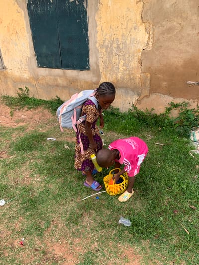 In Kabba, Nigeria, two young children are interacting outdoors next to a weathered, light-colored building with a dark metal-covered window. The child on the left, a girl with braided hair, wears a patterned dress and a light-colored backpack. She is looking down towards the other child. The child on the right, wearing a bright pink t-shirt with white lettering and yellow shoes, is bending over a yellow plastic basket, appearing to be placing items inside. The ground is covered with green grass and patches of dry earth, with some scattered debris including plastic bags and wrappers.