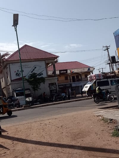 This image captures a street scene in Pantame, Nigeria. In the foreground, a dirt road with shadows suggests the time of day. A paved sidewalk with some overgrown vegetation borders the road. On the sidewalk, a person is riding a motorcycle. Across the street, a white van is loaded with goods on its roof. To the left of the van, a yellow tricycle is parked.
The background features buildings with red-tiled roofs. One building has a large billboard advertising an "Infinix HOT 60 Pro+" smartphone. Another building is adorned with banners and signage. Several people are visible on the sidewalk in front of the buildings, some appearing to be shoppers or pedestrians. Electrical poles and overhead wires are visible against a clear sky. The overall impression is a bustling commercial area with typical street life and transportation.