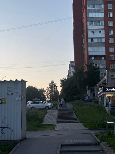 The image depicts an urban street scene in Izhevsk, Russia, under a clear, pale blue sky transitioning to a light orange-yellow hue, indicative of dusk or dawn. Numerous power lines are visible crossing the sky.
On the far left, a corrugated metal utility box or kiosk is present, covered in multiple faded and torn posters primarily in Russian. discernible texts include "РАБОТА" (Work), "СРОЧНО ТРЕБУЕТСЯ" (Urgently Required) with associated phone numbers (e.g., 8-922-517-04-94, 8-912-856-11-94, 8-912-441-31-51), "МАРКЕ ПИТАНИЕ ГАЛАНТ" (Galant Food Market), and "СТРОИТЕЛЬСТВО И РЕМОНТ" (Construction and Repair). Dark graffiti is present on the lower left of the box.
A paved asphalt path leads from the lower left foreground, curving towards the center and connecting to a series of concrete steps. To the right of this path, a wider set of concrete stairs ascends, bordered by a grassy slope on its right. Further up the slope, a blue sign with the "ПОЧТА РОССИИ" (Russian Post) emblem is visible on a pole, near a birch tree.
In the midground, several parked vehicles are present, including a white station wagon equipped with a black roof box. Beyond the parked cars and stairs, various trees with green foliage are visible. In the background right, multiple blue road signs with white arrows indicate directional turns (left, straight, right), alongside a yellow diamond sign signifying a priority road.
The right side of the image is dominated by a tall, multi-story apartment building constructed of red brick with grey concrete panels between window levels. The building features numerous windows, some with enclosed balconies, and external air conditioning units. At its base, a commercial frontage with illuminated windows displays a prominent red sign with white outlining, bearing the Cyrillic text "Хлеб" (Bread).
Several individuals are present: a person in a white top and light pants walks up the steps towards the "Хлеб" store on the far right. Another person in dark clothing ascends stairs in the midground. On the lower set of steps to the left, a woman in a blue top and light pants descends while carrying a bag, followed by another person in dark attire. Other multi-story buildings are visible in the distance to the left of the main brick structure. The ground consists of asphalt, paving stones, and areas of green grass.