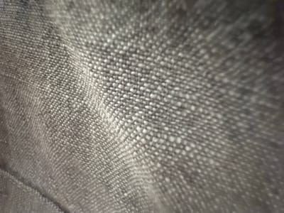 This media file is a close-up, abstract shot of a textured gray fabric. The fabric appears to be woven, with a visible pattern of interlacing threads creating a slightly bumpy surface. Light catches the raised threads, creating bright highlights and casting subtle shadows, which accentuates the texture and gives the fabric a sense of depth. The focus is shallow, with the foreground slightly blurred and the background progressively out of focus, drawing the viewer's attention to the intricate details of the weave in the central and right portions of the image. There are no people, specific objects, or clear scene elements visible. The image is a study in texture and light on a neutral-colored textile. The context provided of "Belgrade, Serbia" does not offer any visual cues within the image itself to determine the specific location or time of day.