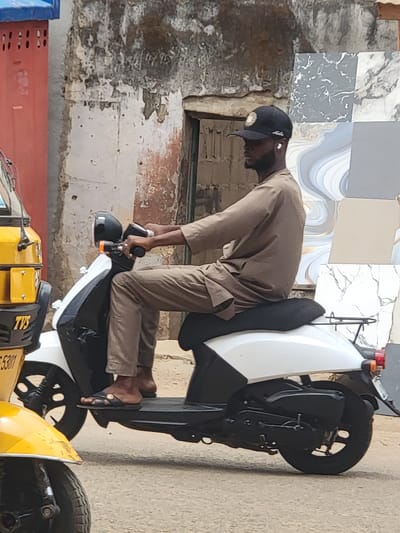 A man wearing a brown shirt, trousers, cap, and sandals is sitting on a white and black motorcycle. He is holding the motorcycle's handlebars. The background consists of a building with grey and white tiling on the upper portion and peeling paint on the lower portion. Other parts of a yellow and black motorcycle are visible at the bottom of the frame. The location is Maiburiji, Nigeria.