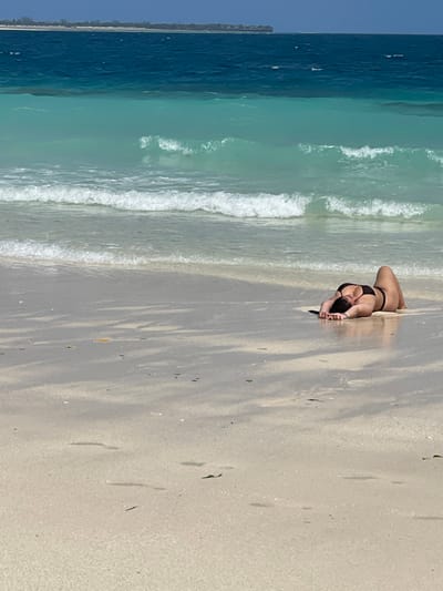 The scene depicts a single individual on a beach in Zanzibar, Tanzania, near Zanzibar City. An adult female is positioned prone on the wet, light-colored sand, close to the water's edge. She is dressed in a black two-piece bikini. Her arms are extended forward, hands resting flat on the sand, and her legs are bent at the knees, slightly elevating her lower body. A light-colored band is visible on her left wrist. The environment includes the Indian Ocean, characterized by layers of clear turquoise water near the shore and deeper blue water extending to the horizon. Several small waves with white crests are breaking on the sandy beach. A low-lying landmass is discernible on the distant horizon under a clear sky. The immediate sand displays subtle patterns and scattered fragments of organic debris.