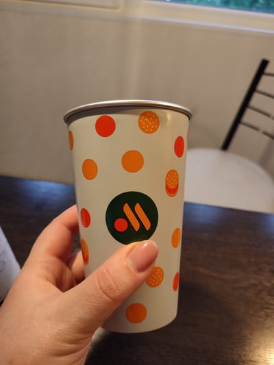 A person's hand with a light pink nail polish is holding a white paper cup with an orange and white dotted pattern and images of hamburgers. The cup has a green circular logo with an orange icon inside. The background is a dark wooden table and a blurred background suggesting an indoor setting. The image was taken in Vsevolozhsk, Russia.
