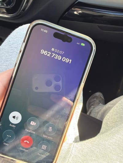 The scene is located within the interior of a vehicle in Vila Real, Portugal. A person holds an iPhone with a clear protective case featuring an orange/gold border. The phone's screen displays an active call interface at 11:46, showing a call duration of 02:07 to the number 962 739 091. The phone's battery is at 77% and appears to be charging, indicated by a lightning bolt icon. Call options in Portuguese, including "Áudio," "FaceTime," "Sem som" (Mute), "Mais" (More), "Desligar" (End Call), and "Teclado" (Keypad), are visible at the bottom of the screen. A distinct reflection of another smartphone with a triple-camera lens array and a hand is visible on the screen's surface. The person holding the phone is wearing light grey casual trousers, which are visible on the lap. A light-colored sneaker, possibly white or light grey, with a dark sole, is partially visible on the vehicle's dark floor mat to the right. The vehicle's interior features dark, glossy surfaces with chrome accents in the background, illuminated by bright sunlight entering from the right side of the frame.