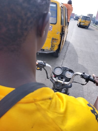 The image depicts a first-person perspective from the back of an individual operating a motorcycle on a multi-lane asphalt road in Alaiyabiagba, Nigeria. The motorcycle operator, whose dark-skinned head and right shoulder are visible, wears a bright yellow shirt with a black strap traversing the right shoulder. Their right hand is positioned on the motorcycle's right handlebar, which features control switches. The motorcycle's instrument panel, including two circular gauges, is visible between the chrome handlebars.
Immediately to the right of the motorcycle, a yellow commercial minibus (commonly known as a "danfo" in Nigeria) is either stopped or moving slowly. The minibus exhibits significant wear, including visible damage to its rear bumper and body panels. Its side sliding door is open, and an adult male, clad in an orange shirt, is partially visible, leaning out or in through the open doorway, holding onto the door frame or roof.
Further down the road, several other vehicles are present, including at least one additional yellow minibus and multiple larger trucks, suggesting moderate to heavy traffic conditions. The sky above is light, indicating an outdoor, daytime scene.