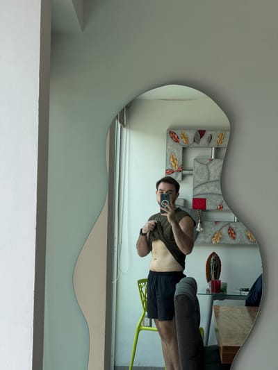 A man is taking a selfie in a mirror. The mirror has an irregular shape with decorative elements around it. The man is wearing a dark green t-shirt, black shorts, and a fitness tracker on his wrist. He is holding a smartphone up to take the photo. In the background, a table with various items is visible, including what appears to be a statue, a red object, and a beaded necklace. A green chair is also in the frame. The wall and room have a modern design with multi-toned paint. The location in the city of San Bernardino Tlaxcalancingo, Mexico is not evident from the image.