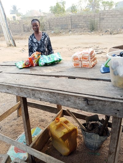 A woman stands behind a weathered wooden table, displaying goods for sale. Bags of chips and stacks of Makowa-branded bread are arranged on the table's surface. Below, a yellow jerrycan, a bucket filled with various objects, and a small shovel rest on the ground beneath the table. In the background, a wall and sparse vegetation are visible. A utility pole stands to the side of the scene.