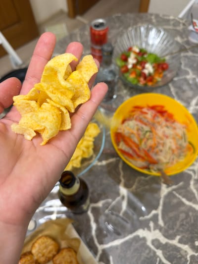 A close-up, over-the-shoulder shot shows a person's hand holding a pile of potato chips. The chips are golden yellow with visible seasoning. In the background, out of focus, is a table with various food items. These include a glass bowl of salad with lettuce, tomatoes, and cheese, a yellow bowl with what appears to be a noodle salad with shredded carrots and red peppers, a bottle of beer, and a red can. The table surface has a marble-like pattern. The scene appears to be indoors. There is no indication of the city of Sochi, Russia in the image.