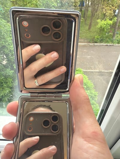 A hand holds an open, hinged compact mirror reflecting a smartphone. The smartphone has a triple-lens camera. The hand's middle finger features a ring. The background, visible through the compact mirror and behind the hand, shows greenery and a paved area, likely an outdoor environment, possibly a park or urban area. The background is also visible through a window frame. The reflection implies the person holding the mirror is taking a picture of themself and their phone. The scene appears to be indoors, given the window frame.