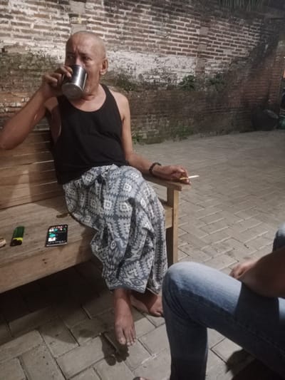 An adult male, bald, with a tan complexion, is seated on a wooden bench in what appears to be an outdoor area in Kecamatan Pati, Indonesia. He is wearing a black tank top and a patterned sarong. The man is drinking from a metal cup held in his right hand and is holding a lit cigarette in his left hand. Next to him on the bench are a green lighter and a mobile phone. The background consists of a brick wall and a paved ground. Another person, wearing blue jeans, is partially visible in the foreground, to the right of the frame. The scene suggests a casual, possibly late-evening setting.