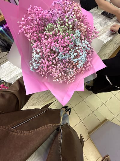 The image shows a bouquet of colorful baby's breath flowers wrapped in bright pink paper. The bouquet is positioned on a table, with a person's legs and feet visible in the foreground. The person is wearing a brown leather jacket and blue jeans, and black pointed-toe heels. In the background, another person's hands are visible on the table, near a calculator and a small red object. The environment appears to be an indoor setting with tiled flooring. This scene is located in the city of Khimki, Russia.