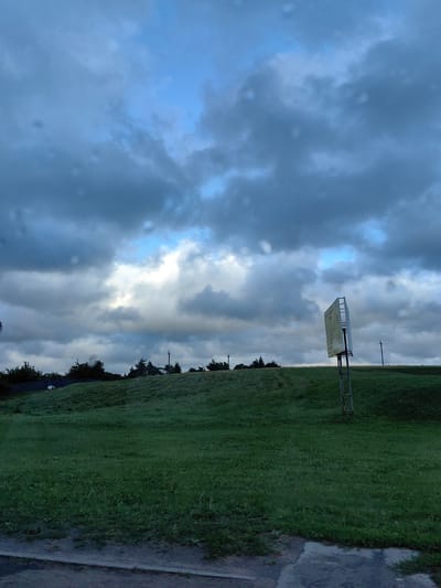 The image depicts a grassy hill under a dramatic, cloudy sky with patches of blue. On the right side of the hill, a tall, metal signpost holds a rectangular sign with yellow and white lettering. The sign is tilted and appears weathered. The hill is covered in lush green grass, and in the distance, a line of trees and utility poles are visible. The lower portion of the image shows a paved surface, possibly a road or sidewalk, with a reflective puddle. The overall atmosphere is overcast and possibly damp, with subtle indications of rain or moisture on the camera lens. The scene appears to be an outdoor landscape, likely in a rural or suburban area, possibly near a road. There are no people visible in the image. Based on the provided context, the location is identified as Фаніпальскі сельскі Савет, Belarus.