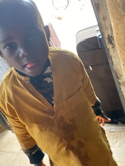 In Bukuru, Nigeria, a young boy wearing a yellow shirt with a camouflage pattern underneath sticks his tongue out at the camera, his face partially obscured by shadow. He stands in a doorway, with luggage visible in the background. The scene appears to be indoors, with natural light streaming in from the right side of the frame. The wall to the right is rough and textured, suggesting it may be made of mud or concrete.