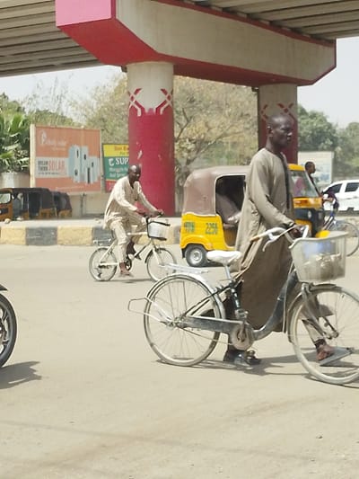 The image displays an outdoor road scene in Fagge A, Nigeria, beneath a concrete structure featuring red horizontal beams and white pillars adorned with a white geometric pattern. In the foreground, a dark-skinned man wearing a light brown tunic walks beside a dark-framed bicycle with a white seat and a clear front basket containing light-colored items. His right foot is on the ground, and his left is on the pedal. To his left and slightly behind him, another dark-skinned man in a light beige patterned tunic is actively pedaling a smaller, white-framed bicycle, also equipped with a front basket. A third person on a bicycle is partially visible further in the background to the right. Two yellow and brown motorized tricycles (Keke Napep) are present; one is in the mid-ground behind the smaller bicycle, displaying "2256" on its rear, and another is further back on the right. A white car is partially visible in the distant right background. In the bottom-left foreground, a black motorcycle wheel is visible. The road surface is light grey asphalt. On the left, a low concrete barrier with yellow and black stripes separates the road. A large billboard for "Fidelity solar" with "DREAM THINK SOLAR" and images of solar panels is prominently displayed on the left. Sparse trees and vegetation are visible in the background, and the scene is illuminated by bright daylight, casting shadows on the road surface.