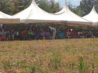 The image shows a gathering of people in Lwandeti, Kenya (latitude: 0.554, longitude: 34.829). The people are seated under several white tents, facing the viewer. There are numerous people seated in rows under the tents, with some individuals standing. Many of the people are wearing brightly colored clothing. The tents appear to be arranged in a line, with a number of trees visible in the background. The ground in front of the tents is covered in dry grass and other vegetation. There is a sign on one of the tents that reads "Tel: 0744 643 622."