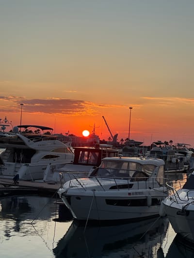 The image shows a marina in Sochi, Russia, during sunset. The sun is setting on the horizon, casting a warm orange glow across the sky and clouds. Several yachts are docked in the marina, their white hulls reflecting in the calm water. A crane and other harbor structures are visible in the background. The scene is peaceful and serene, with no people visible in the frame.