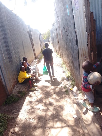 The image depicts a narrow, unpaved dirt alleyway within an informal settlement, likely a slum such as Kibera or Mathare, in Nairobi, Kenya. The alley is bordered on both sides by structures primarily constructed from corrugated metal sheets, exhibiting signs of wear and rust. A shallow concrete drainage channel runs along the right side of the path, with small patches of green vegetation growing at the bases of the structures. Overhead, harsh daylight illuminates the scene, casting dappled shadows on the ground and creating a bright overexposure at the far end of the alley.

On the left side of the frame, four individuals are seated on the ground or a low ledge against the corrugated metal wall. The most prominent of this group wears a bright yellow long-sleeved top and dark pants, with light green slip-on shoes. Others in the group wear dark and light clothing, partially obscured. In the mid-ground, centered in the path and facing away from the viewer, an individual in a dark t-shirt and dark pants walks away, carrying a bright green plastic bag in their left hand. On the right side of the frame, a young child, appearing to be a toddler, stands facing left. The child wears a striped long-sleeved shirt (red, blue, white), light-colored pants, and light green slip-on shoes. They hold a light-colored, possibly worn or deflated, ball in both hands and appear to be touching the corrugated wall with their right hand. No direct interpersonal interaction is observed between the distinct groups of individuals.