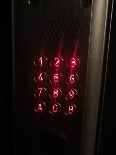 A close-up, low-light photograph displays a metallic access control keypad, likely situated at an entrance point within the city of Paris, France. The keypad is constructed from a dark, possibly brushed aluminum or similar metal, secured by two visible screws at the top corners. Centered above the input buttons is a rectangular grille, perforated with multiple small circular holes, consistent with a speaker or microphone. The keypad comprises twelve circular, illuminated buttons arranged in four rows of three columns. These buttons display red alphanumeric characters: '1', '2', '3' in the top row; '4', '5', '6' in the second; '7', '8', '9' in the third; and 'A', '0', 'B' in the bottom row. Distinct vertical streaks of red light extend across the button surfaces, indicating reflections from the internal LED illumination or ambient light. The surrounding area is obscured by darkness, suggesting a dimly lit indoor environment or nighttime conditions. No individuals or active interactions are visible in the scene.