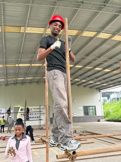 An outdoor scene at the Glodon Construction Industrialization College in Jiangjin, China, features a young Black man, wearing a red hard hat and work gloves, giving a thumbs-up while standing on scaffolding. He is dressed in a black t-shirt and gray pants. Behind him, several other individuals in work attire are also on scaffolding. In the foreground, a young Black woman wearing a pink jacket and gray shirt holds some small wooden objects in her gloved hand. The environment consists of a large open-air structure with a metal roof, concrete ground, and scattered construction materials, primarily metal pipes and scaffolding. A sign in the background displays Chinese characters and the Glodon logo. The weather appears to be overcast.