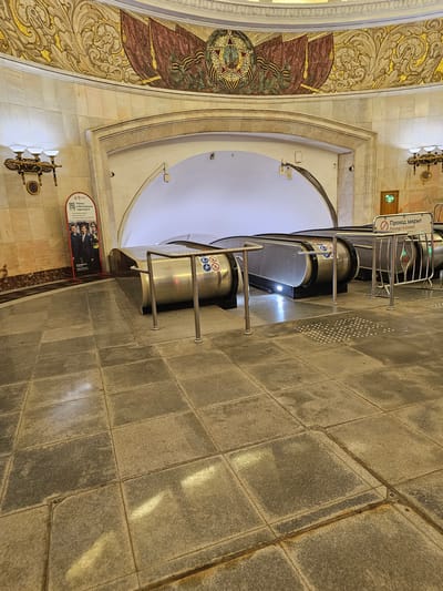 The image shows an interior view of a Moscow Metro station, likely the Komsomolskaya station, given its ornate architectural style with gilded mosaics and marble walls. In the foreground, a gray tiled floor leads towards a pair of escalators. One escalator is to the left and appears to be active with some lights illuminating its base. The escalator to the right is blocked by metal barriers and a sign that reads "Проход закрыт Passage is closed" in Russian and English. To the left of the active escalator, a red standing banner displays information about jobs in Moscow transport, featuring images of uniformed workers. The background features a grand arched entrance with a brightly lit interior beyond, and above the arch, a large mosaic adorns the curved ceiling, depicting a stylized banner and a star. The overall impression is one of grandeur and public transportation infrastructure.