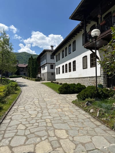 The image shows a view of a stone-paved walkway adjacent to a white building with dark-framed windows. The building has a stone base and a dark wooden roof. The sky is blue with white clouds. Vegetation is present on either side of the walkway, including grass, shrubs, and trees. In the background, there is a mountain and other buildings. Based on the provided information, this could be located in Barziya, Bulgaria.
