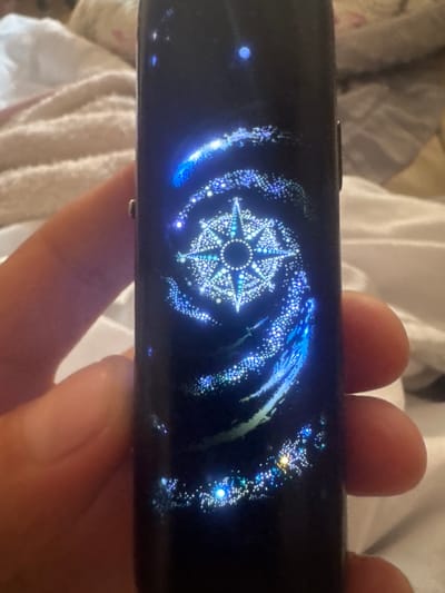 The image shows a close-up of a person's fingers holding a dark, rectangular electronic device. The device has a screen displaying a vibrant, glowing graphic that resembles a starry spiral or a cosmic vortex. At the center of the graphic is a complex, star-like geometric pattern, possibly a stylized compass rose, made up of many small, bright dots. The overall color palette of the screen display is dominated by blues, whites, and some hints of green and yellow, creating a sense of depth and ethereal beauty. The background is blurred and appears to be a white fabric, possibly a towel or bedding, suggesting an indoor setting. The lighting on the device screen is bright, illuminating the surrounding fingers and the edge of the device. No visible text is present. The activity appears to be the simple act of holding and displaying the device. The focus is on the illuminated screen and the graphic it displays.