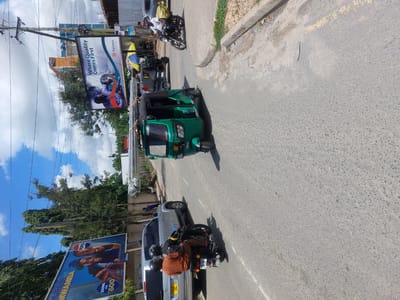 This image depicts a bustling street scene in Dar es-Salaam, Tanzania, under a bright, partly cloudy sky during daylight hours. The scene is filled with vehicles and urban infrastructure.
The main subjects include various modes of transport: a prominent green three-wheeled auto-rickshaw (tuk-tuk or bajaj) in the middle of the road, a silver SUV or car with the license plate "T513 EGL", and at least two motorcycles. One motorcycle is positioned next to the silver car, with its rider wearing an orange shirt and a helmet; its license plate is "MC507 EJZ". Another motorcycle is visible further down the street.
Along the roadside, utility poles with numerous overhead wires are present. Two large advertising billboards are highly visible. One billboard, advertising "Salama," features the slogan "Where Quality Comes First" and shows two individuals. The second billboard promotes "PEPSI" and includes the text "HUPANI MWAMBA" and "350ML KWA 600," likely referring to a product size and price. Both billboards display the "JCDecaux" logo.
In the background, several buildings are visible, including one labeled "SONI HOUSE" and another with a sign for "PRIS SPA 7467373." Trees and other vegetation are interspersed among the buildings and alongside the road. The road surface is asphalt, and there's a concrete curb separating it from a dirt and grassy area. The scene captures the daily activity and traffic of a lively urban setting.