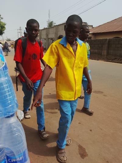 The image depicts an outdoor daytime street scene in Akwanga, Nigeria, under an overcast sky. In the foreground, partially visible on the far left, is a stack of clear plastic water bottles with blue caps, bearing the partially visible brand "CWAYS". Three young males walk from left to right on an unpaved, reddish-brown dirt road. The male closest to the viewer, positioned center-right, wears a bright yellow short-sleeved button-up shirt with a blue collar, blue trousers, and brown open-toed sandals. His left arm is bent, with his index finger pointing downwards and slightly forward. A dark necklace is visible around his neck. To his left and slightly behind him, another male wears a red short-sleeved t-shirt with a dark circular logo, blue trousers, and light-colored open-toed sandals, carrying a black backpack. To the right and slightly behind the first male, a third male is partially visible, wearing a light-colored top, blue trousers, and dark open-toed sandals.
In the midground and background, additional individuals are observed walking along the street. Structures include a low brick wall and a building with a corrugated metal roof on the right. On the left, utility poles with wires are visible, along with concrete buildings. A palm tree and other vegetation are present. A tall cellular tower or radio mast stands against the light grey sky in the far background. The ground shows irregular patches and darker stains.