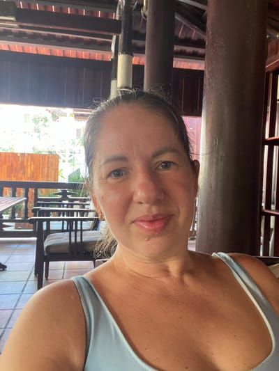 A woman is taking a selfie in an outdoor setting. She has light skin and is wearing a light blue tank top. Her hair is pulled back, and she is smiling at the camera. In the background, there are wooden tables and chairs, suggesting she is at a cafe or restaurant. The architecture appears to be a covered patio with wooden beams and a tiled floor. The scene is lit by natural sunlight coming from the left. The specific location in Phu Thuy Ward, Vietnam, is not visually identifiable from the provided image.