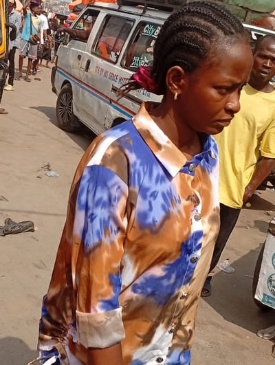 The image depicts a bustling street scene in Fegge, Nigeria, under bright daylight. In the foreground, a young adult female with dark skin, styled in braided cornrows pulled back with a reddish-pink scrunchie, is captured from the chest up, angled slightly away from the viewer. She wears a long-sleeved, collared button-up shirt with a distinct blue, brown, and white tie-dye or camouflage pattern, and a small gold-colored hoop earring is visible in her right ear. Her expression is neutral, and she gazes towards the right side of the frame.
To her immediate right and slightly behind, a man with dark skin wearing a plain yellow short-sleeved t-shirt is partially visible, looking forward. In the midground, a white passenger van, marked with "City Bus" on its rear window and "ITS BY HIS GRACE MASTER TRANSITION LTD." along its side, occupies a significant portion of the street. The van features custom black and chrome wheel rims, and a person's arm extends out from the driver's side window. Beyond the van, several individuals are present, some walking, including a man carrying an object on his head, suggesting commercial or transit activity. The paved street surface shows some wear and scattered debris, including a dark, irregular object near the foreground subject's left foot. The background features indistinct urban structures under a clear sky.