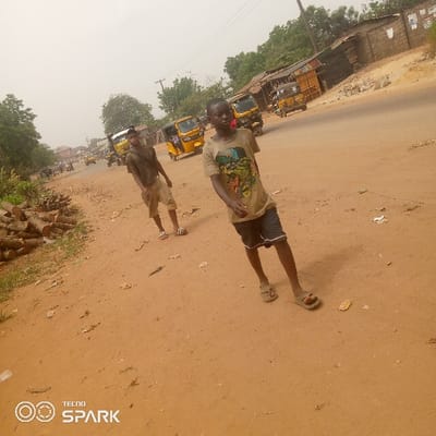 The scene depicts two young males walking on a reddish-brown dirt shoulder adjacent to a main road in Onitsha, Nigeria. The foreground subject, a dark-skinned male, wears a light-colored t-shirt with a graphic design and dark shorts, with light-colored sandals on his feet. He is positioned towards the right side of the frame, looking directly at the camera. Behind and to his left, a second dark-skinned male wears a dark t-shirt and light shorts, with striped sandals. He also looks towards the camera.
To the left of the subjects, a substantial pile of cut logs is stacked on the dirt ground. The main road runs parallel to the dirt shoulder, containing multiple yellow tricycle taxis (Keke Napep) that are either stationary or in motion. Beyond the road, single-story structures, constructed from concrete blocks with some corrugated roofing, line the street. Sparse green trees and bushes are visible in the background and along the left side. The sky appears hazy or overcast. Debris, including small pieces of paper or plastic, is scattered across the dirt ground.