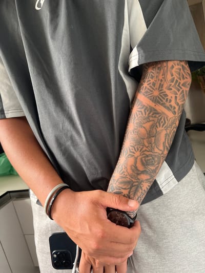A close-up, medium shot shows a person's torso and arm, wearing a gray and white t-shirt and gray sweatpants. The person's left arm is covered in a dark tattoo that extends from the wrist to the elbow. The tattoo features geometric patterns and roses. A watch with a black band is on the person's right wrist, and two bracelets, one light gray and one black, are on the left wrist. A black smartphone is held in the person's right hand. The background is blurred and appears to be an indoor setting, possibly a kitchen or living area. There is no indication of the location being Madrid, Spain.