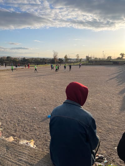 The scene depicts an unpaved outdoor sports field in Fez, Morocco, observed from a low vantage point. In the immediate foreground, a person is seen from behind, wearing a dark blue denim jacket over a red hooded sweatshirt, seated or crouched on uneven, refuse-strewn dirt. Ahead, approximately 20-30 individuals are scattered across the expansive, dusty field. Many are dressed in casual sportswear, with some distinguished by green shirts, indicating participation in an informal football (soccer) game or practice. Long shadows stretch eastward across the field, consistent with late afternoon sunlight. The field's perimeter features sparse vegetation and some wire fencing, with a distant urban landscape of buildings visible against a hazy horizon. The sky is partially cloudy with patches of clear blue.