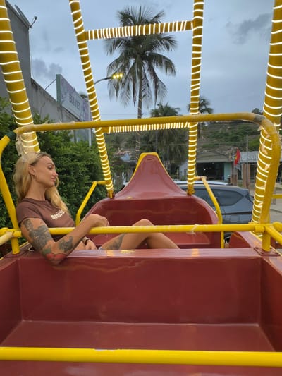 A woman with blonde hair and visible tattoos on her arms and legs sits in a red gondola with yellow trim, illuminated by string lights. She is wearing a brown t-shirt with white lettering. The gondola appears to be part of a Ferris wheel. In the background, palm trees and buildings suggest a tropical location. A sign visible in the background reads "BIA VIET." The scene is set at dusk with a cloudy sky.