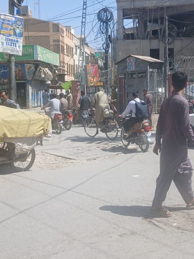 The image depicts a street scene in Quetta City Tehsil, Pakistan, showing a line of people on bicycles and motorcycles traveling down a street. Several buildings are visible in the background with numerous signs advertising businesses, and tangled power lines and barbed wire overhead. A person in sandals is walking along the side of the street in the upper right corner of the frame.