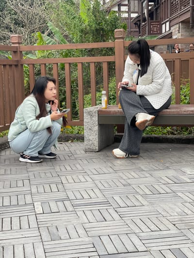 This image depicts an outdoor scene in Shapingba District, China, featuring two adult females on a paved area with traditional architecture in the background.

The ground is composed of rectangular grey paving stones arranged in an alternating basketweave pattern. A dark wooden bench, supported on the left by a grey stone block, is positioned to the right of the frame's center. A wooden fence with horizontal planks and vertical posts extends horizontally across the mid-ground.

On the left, a woman with long dark hair, dressed in a light-colored (possibly light green) jacket, light blue jeans, and black and pink athletic shoes, is in a deep squat. She holds a silver-colored smartphone in her right hand and rests her left hand near her chin, looking towards the left with a focused expression.

On the right, a second woman with dark hair pulled back into a high ponytail, wearing a white quilted jacket, dark grey wide-leg trousers with light polka dots, and white and tan chunky-soled athletic shoes, is seated on the wooden bench. She holds a silver-colored smartphone with both hands, directing her gaze downwards at the screen. A clear plastic bottle containing an orange beverage with a white cap rests on the bench to her left.

Behind the wooden fence, dense green foliage, including broad-leafed plants, is visible. Further in the background, traditional Chinese buildings with dark wooden frames, white walls, and tiled roofs are present. A person in dark clothing is partially visible behind the fence in the upper right background.